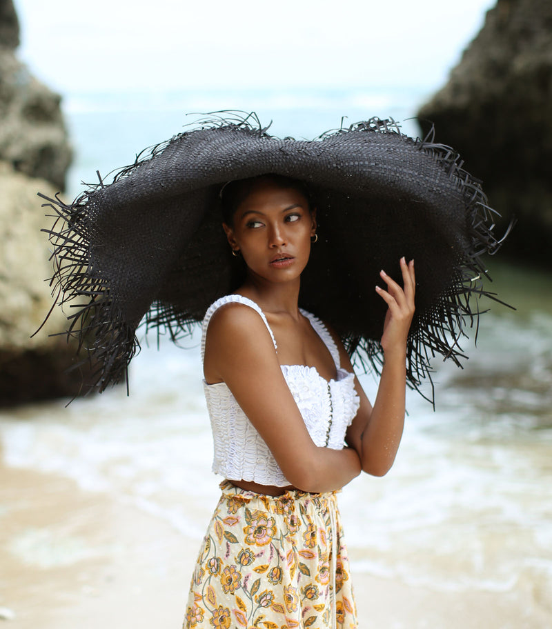 A woman is wearing BrunnaCo’s Amora Handwoven Frayed Oversized Straw Hat in black made by artisans in Bali. This tropical island hat is made for a perfect holiday vacation and coastal getaways. The photograph is taken by Faruq Adib’s team, Matarasa in Bingin Beach, Bali.
