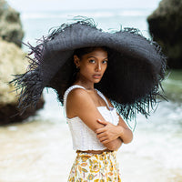 A woman is wearing BrunnaCo’s Amora Handwoven Frayed Oversized Straw Hat in black made by artisans in Bali. This tropical island hat is made for a perfect holiday vacation and coastal getaways. The photograph is taken by Faruq Adib’s team, Matarasa in Bingin Beach, Bali.