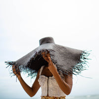 A woman is wearing BrunnaCo’s Amora Handwoven Frayed Oversized Straw Hat in black made by artisans in Bali. This tropical island hat is made for a perfect holiday vacation and coastal getaways. The photograph is taken by Faruq Adib’s team, Matarasa in Bingin Beach, Bali.