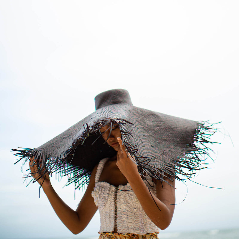A woman is wearing BrunnaCo’s Amora Handwoven Frayed Oversized Straw Hat in black made by artisans in Bali. This tropical island hat is made for a perfect holiday vacation and coastal getaways. The photograph is taken by Faruq Adib’s team, Matarasa in Bingin Beach, Bali.