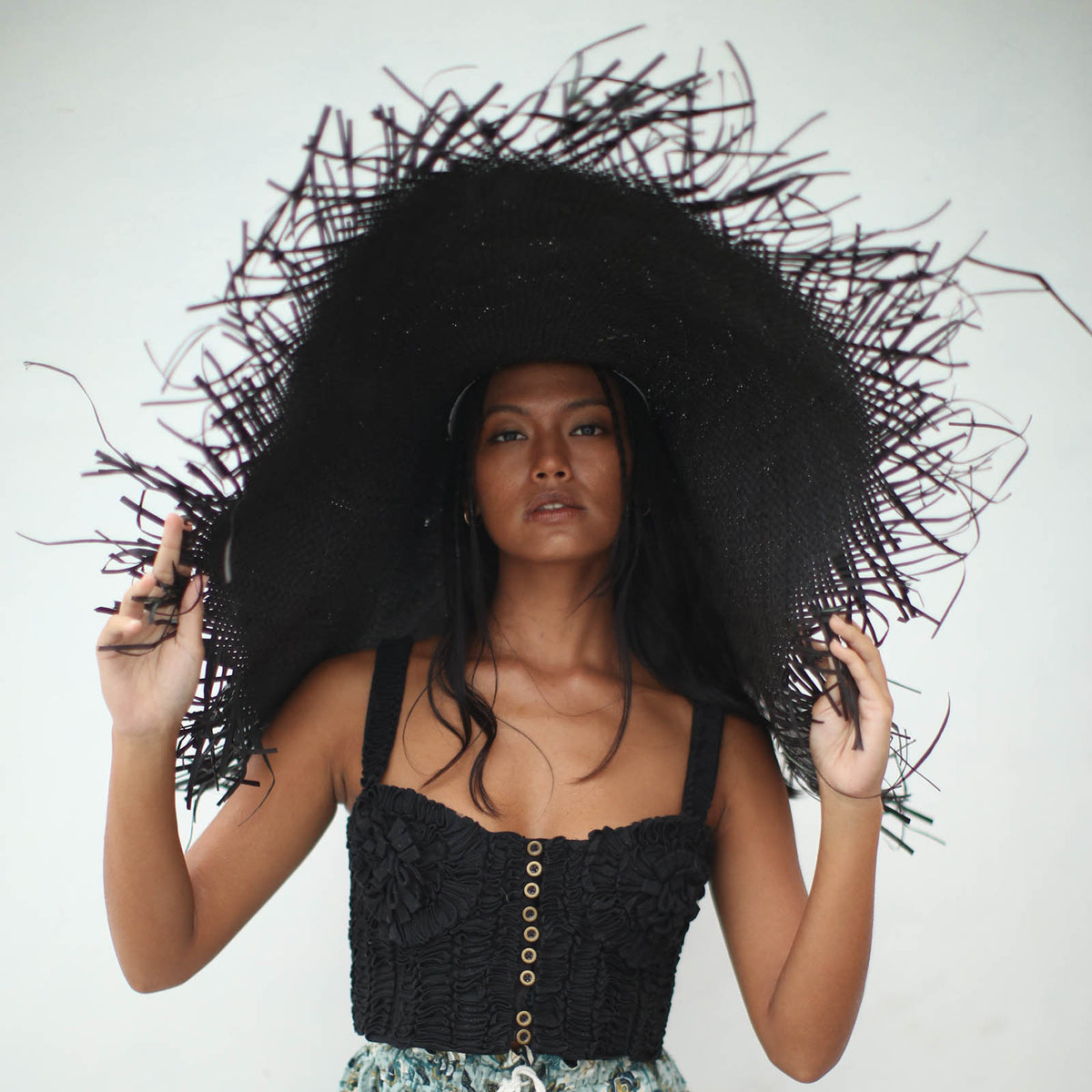 A woman is wearing BrunnaCo’s Amora Handwoven Frayed Oversized Straw Hat in black made by artisans in Bali. This tropical island hat is made for a perfect holiday vacation and coastal getaways. The photograph is taken by Faruq Adib’s team, Matarasa in Bingin Beach, Bali.