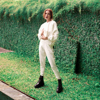A woman stands outdoors in front of a green ivy-covered wall, wearing a loose white sweatshirt and matching white jogger pants with black combat boots.