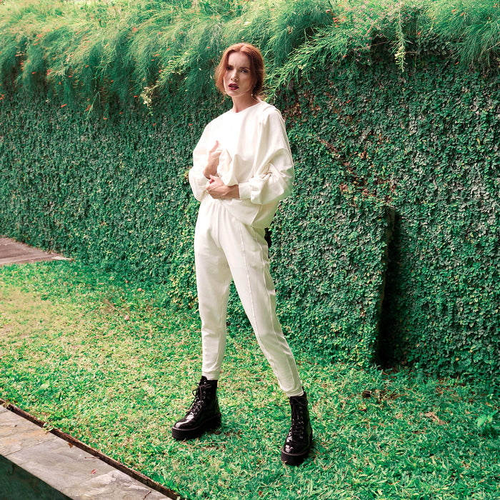 A woman stands outdoors in front of a green ivy-covered wall, wearing a loose white sweatshirt and matching white jogger pants with black combat boots.