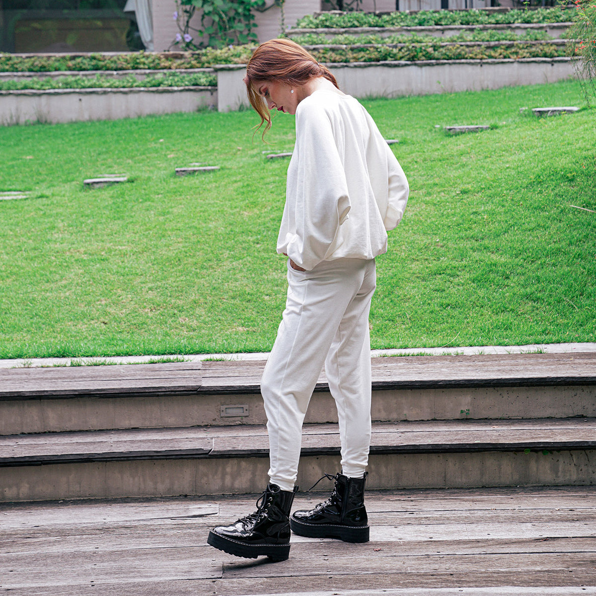 A person standing outdoors on wooden steps in front of a grassy park area, wearing an oversized white top and matching white pants with black lace-up boots.