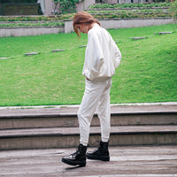 A person standing outdoors on wooden steps in front of a grassy park area, wearing an oversized white top and matching white pants with black lace-up boots.