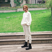 A model stands on outdoor wooden steps in front of a green lawn, wearing a loose white long-sleeve top and white jogger pants with black platform combat boots.