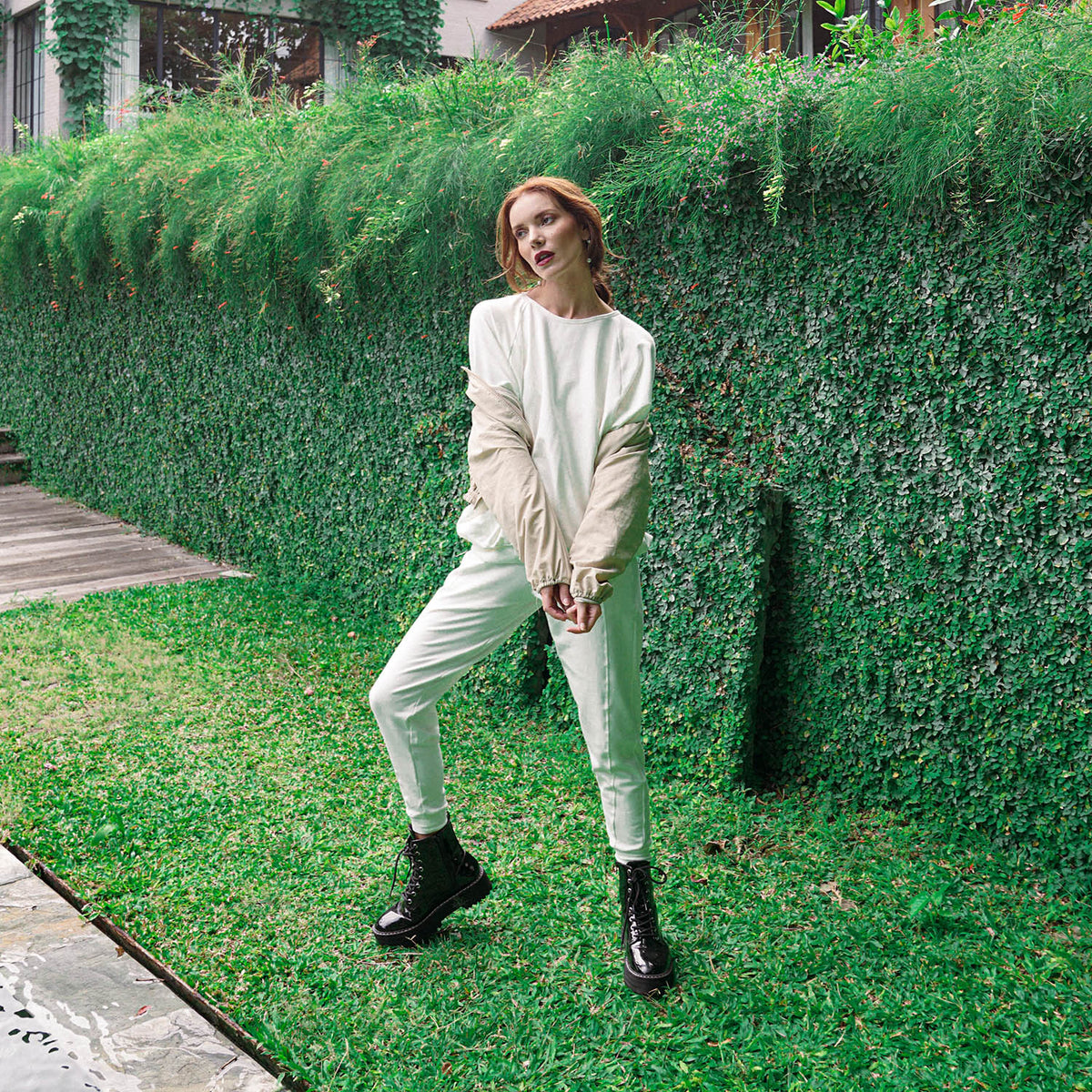 A woman posing outdoors in front of a lush green hedge, wearing a cream-colored long-sleeve top and matching cream pants, paired with black lace-up boots.
