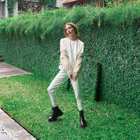A woman posing outdoors in front of a lush green hedge, wearing a cream-colored long-sleeve top and matching cream pants, paired with black lace-up boots.