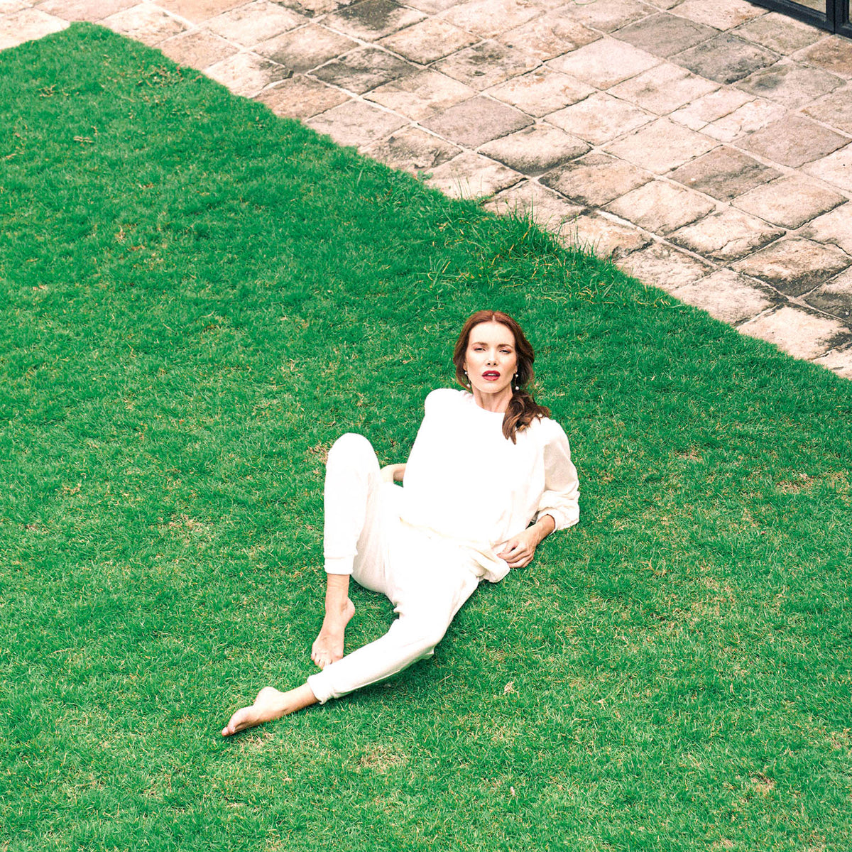A woman wearing a matching cream-colored two-piece outfit sits on a green lawn next to a stone patio.