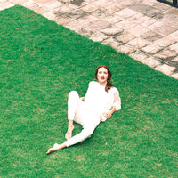 A woman wearing a matching cream-colored two-piece outfit sits on a green lawn next to a stone patio.