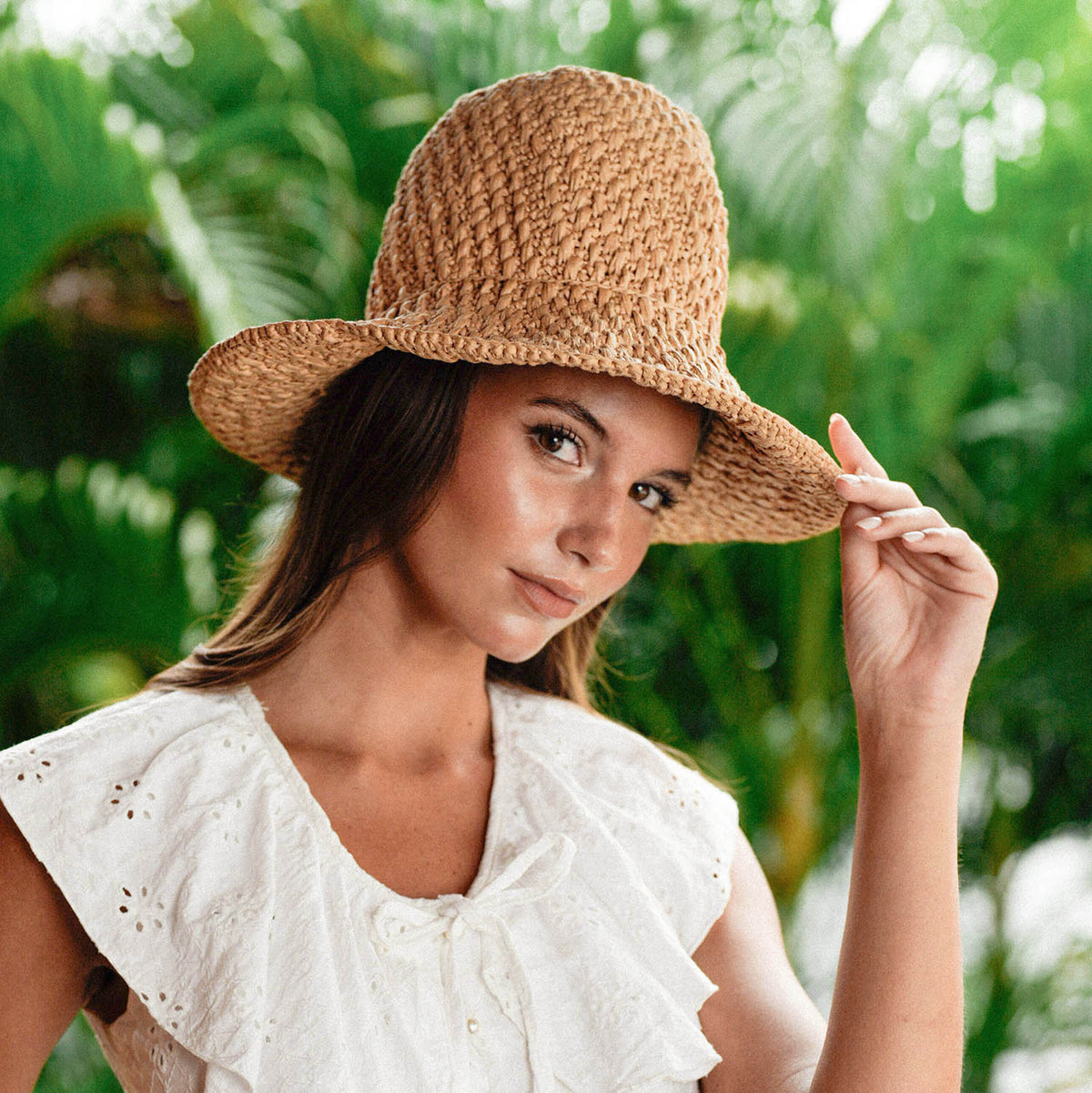 A Woman, Rachelle Van Waardhuizen is wearing BrunnaCo's artisanal Nina raffia straw hat and white Padar embroidery dress with a blurred green background of a lush tropical jungle in Bali