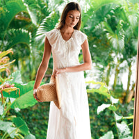 Woman in a white embroidered vacation dress holding a straw hat amidst green foliage