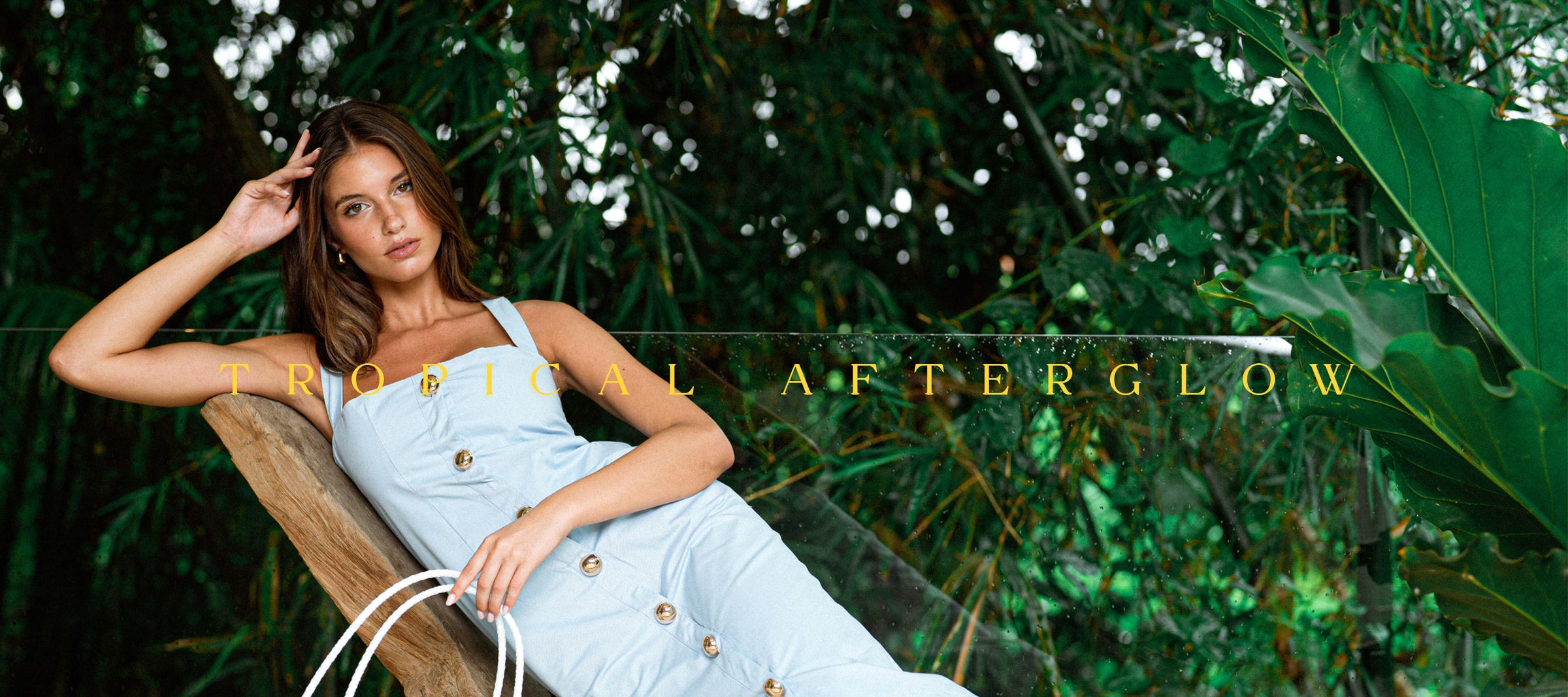 Rachel Van Waanderhuizen reclines on a wooden lounge chair outdoors among lush green tropical foliage, wearing a light blue sleeveless jumpsuit with gold-tone buttons. The text 'TROPICAL AFTERGLOW' is overlaid across the center.