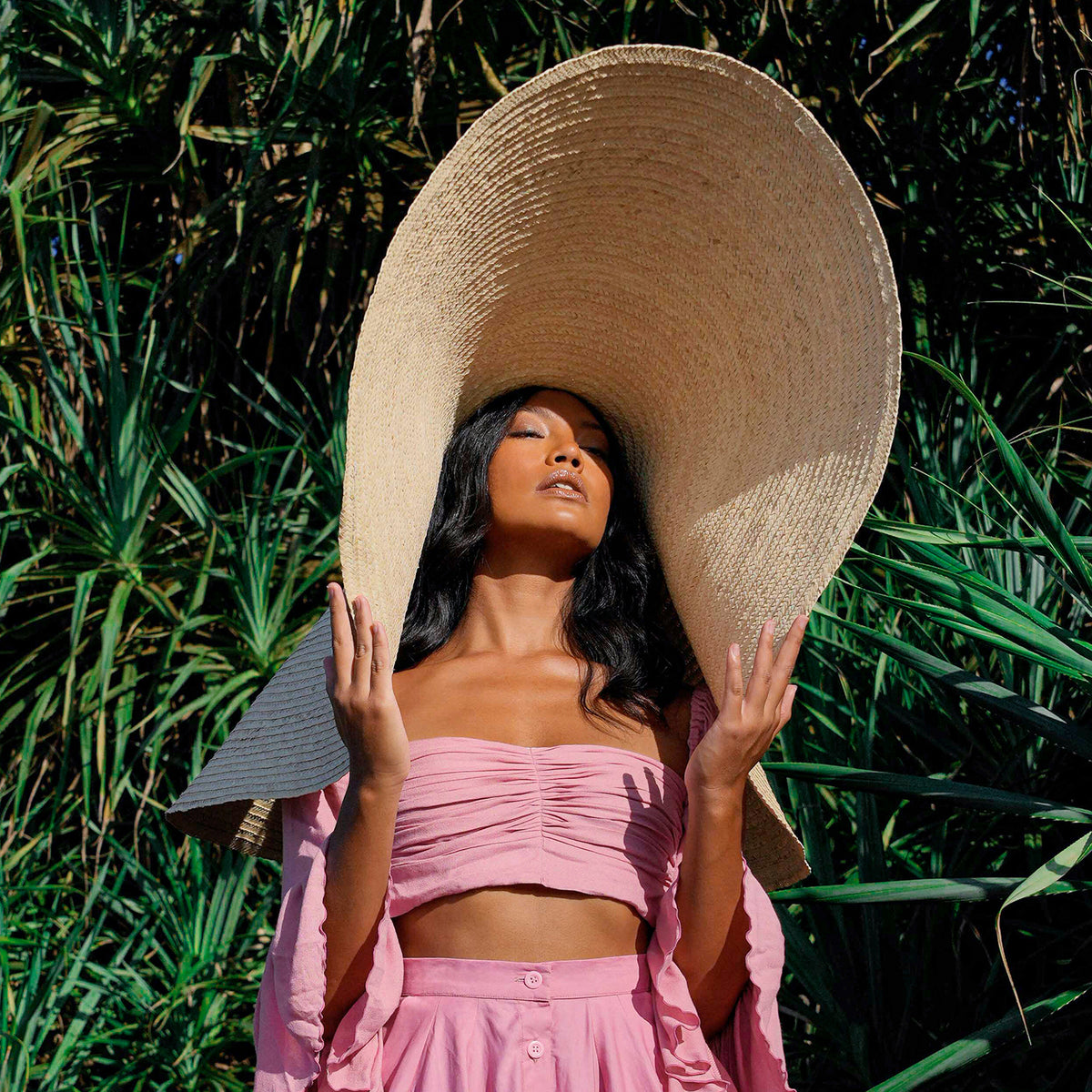 Inka Williams stands on a sun-drenched beach in Bali, wearing an oversized Solana Straw Hat that casts soft shadows across her face. The golden sand, gentle waves, and clear blue sky frame the effortless elegance of her look.