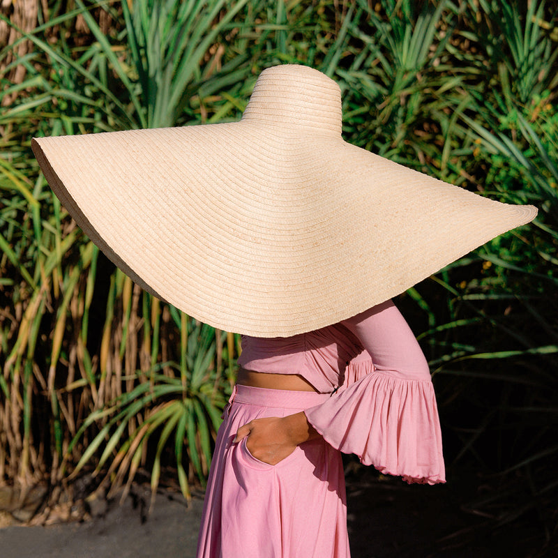 Inka Williams stands on a sun-drenched beach in Bali, wearing an oversized Solana Straw Hat that casts soft shadows across her face. The golden sand, gentle waves, and clear blue sky frame the effortless elegance of her look.