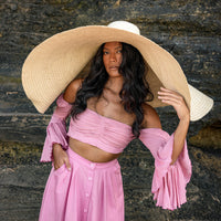 Inka Williams stands on a sun-drenched beach in Bali, wearing an oversized Solana Straw Hat that casts soft shadows across her face. The golden sand, gentle waves, and clear blue sky frame the effortless elegance of her look.