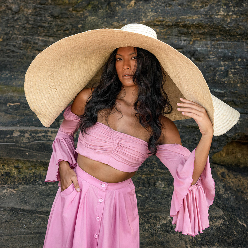 Inka Williams stands on a sun-drenched beach in Bali, wearing an oversized Solana Straw Hat that casts soft shadows across her face. The golden sand, gentle waves, and clear blue sky frame the effortless elegance of her look.