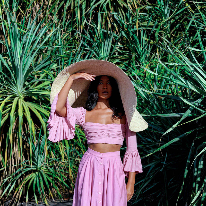 Inka Williams stands on a sun-drenched beach in Bali, wearing an oversized Solana Straw Hat that casts soft shadows across her face. The golden sand, gentle waves, and clear blue sky frame the effortless elegance of her look.