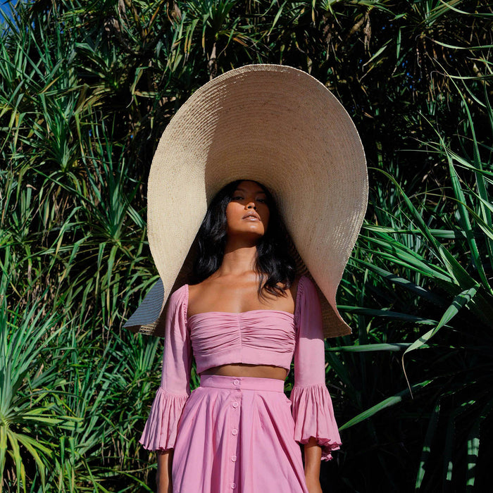 Inka Williams stands on a sun-drenched beach in Bali, wearing an oversized Solana Straw Hat that casts soft shadows across her face. The golden sand, gentle waves, and clear blue sky frame the effortless elegance of her look.