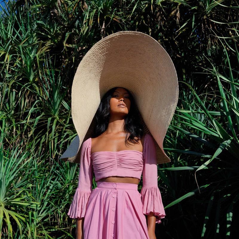 Inka Williams stands on a sun-drenched beach in Bali, wearing an oversized Solana Straw Hat that casts soft shadows across her face. The golden sand, gentle waves, and clear blue sky frame the effortless elegance of her look.