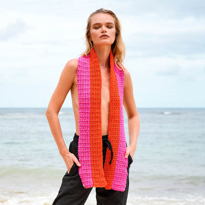 A woman standing on a beach wearing a chunky crocheted wrap scarf with vertical stripes of bright orange and hot pink.