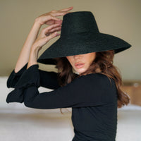 A woman wearing The Riri Jute Woven Straw Hat in Black at a beach in Bali, the hat was made sustainability by artisans in Bali